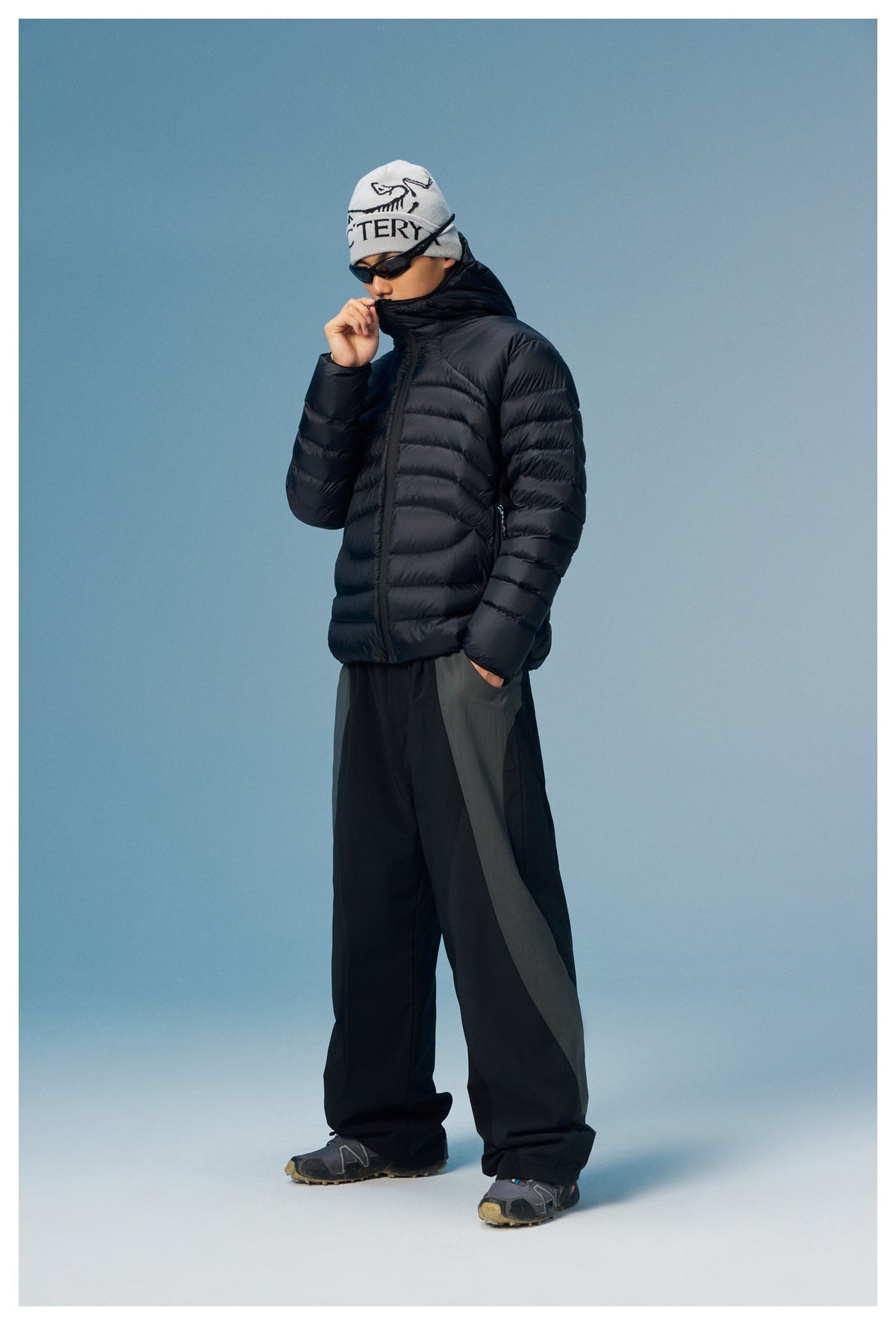 Full-length model wearing a black quilted puffer jacket, loose black trousers, sunglasses, and a light beanie.