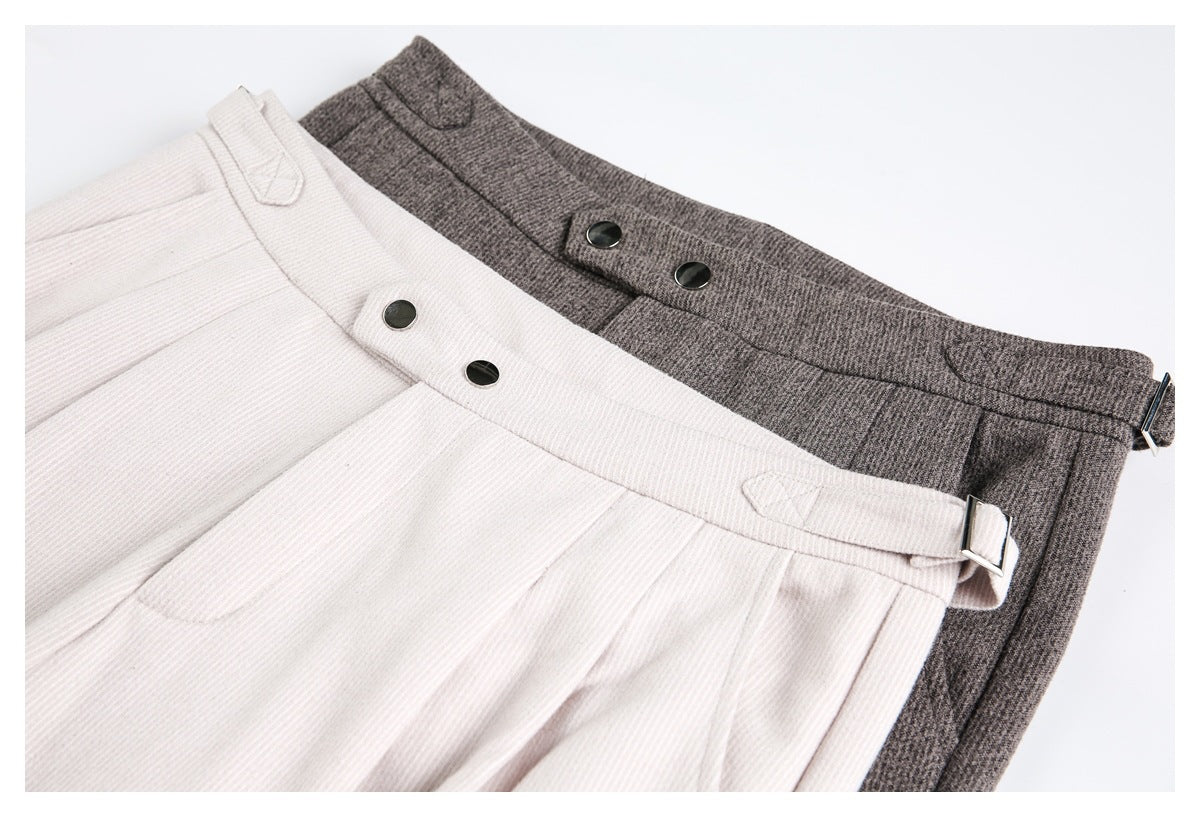 Close-up of ivory and charcoal trousers showing pleated waistbands, dark buttons, and adjustable side tabs.