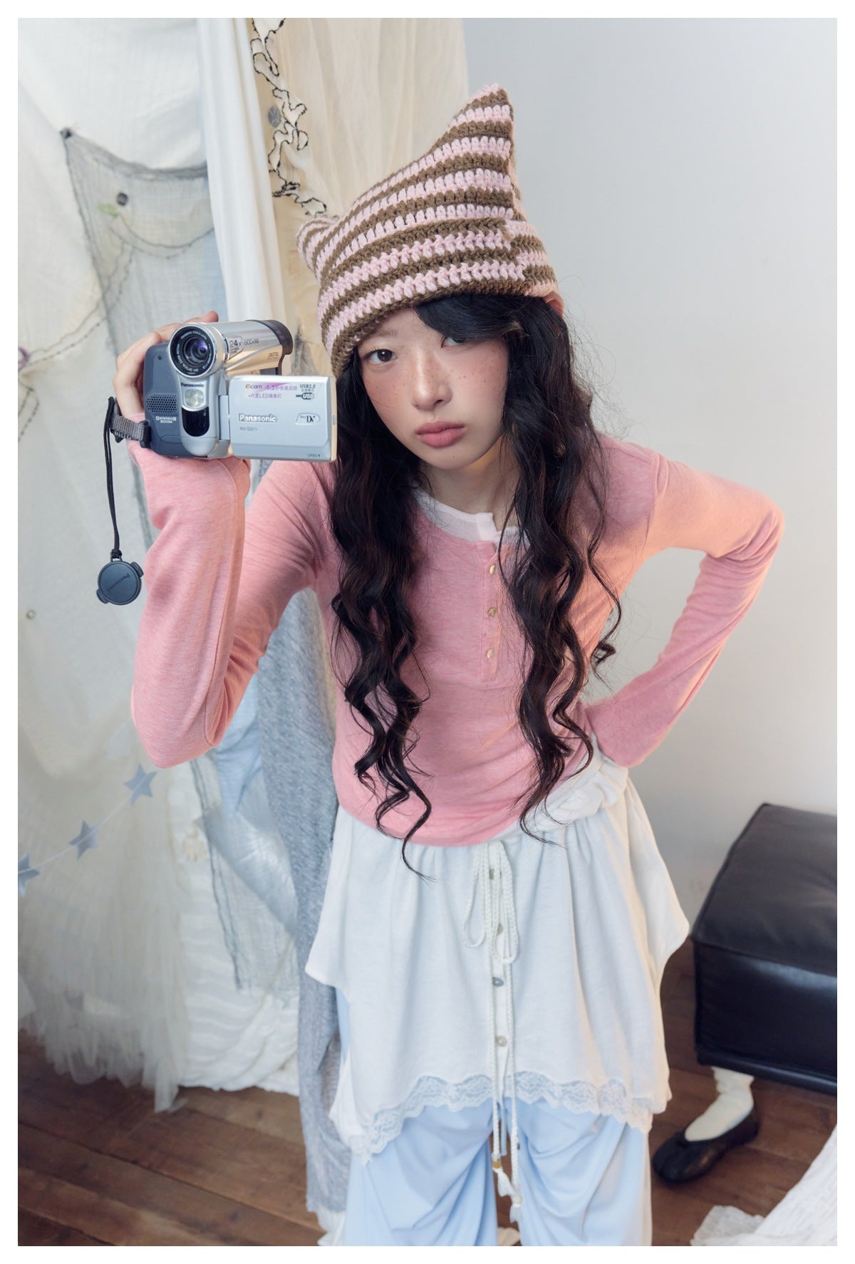 Model wearing a pink long-sleeve henley with a white lace-trimmed wrap skirt and pale blue trousers.