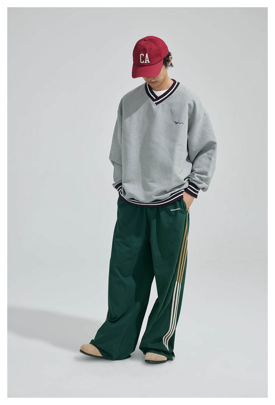 Model looking down wearing a gray V-neck striped-trim sweatshirt, red cap and wide green striped pants