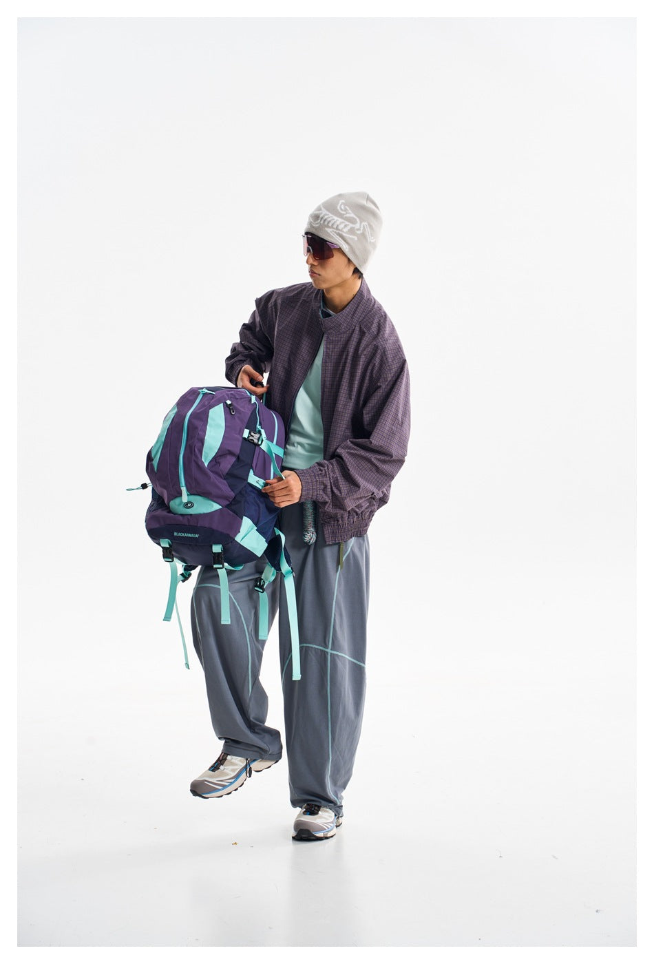 Model wearing a dark purple checked jacket and gray piped trousers while holding a purple and turquoise backpack.