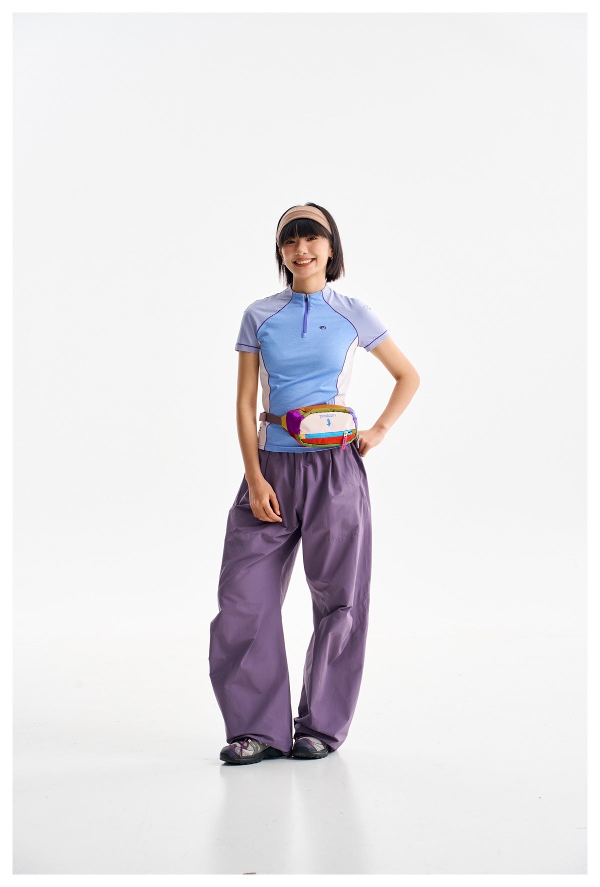 Model wearing a blue and lavender short-sleeve zip top, loose purple pants and a multicolored waist bag.