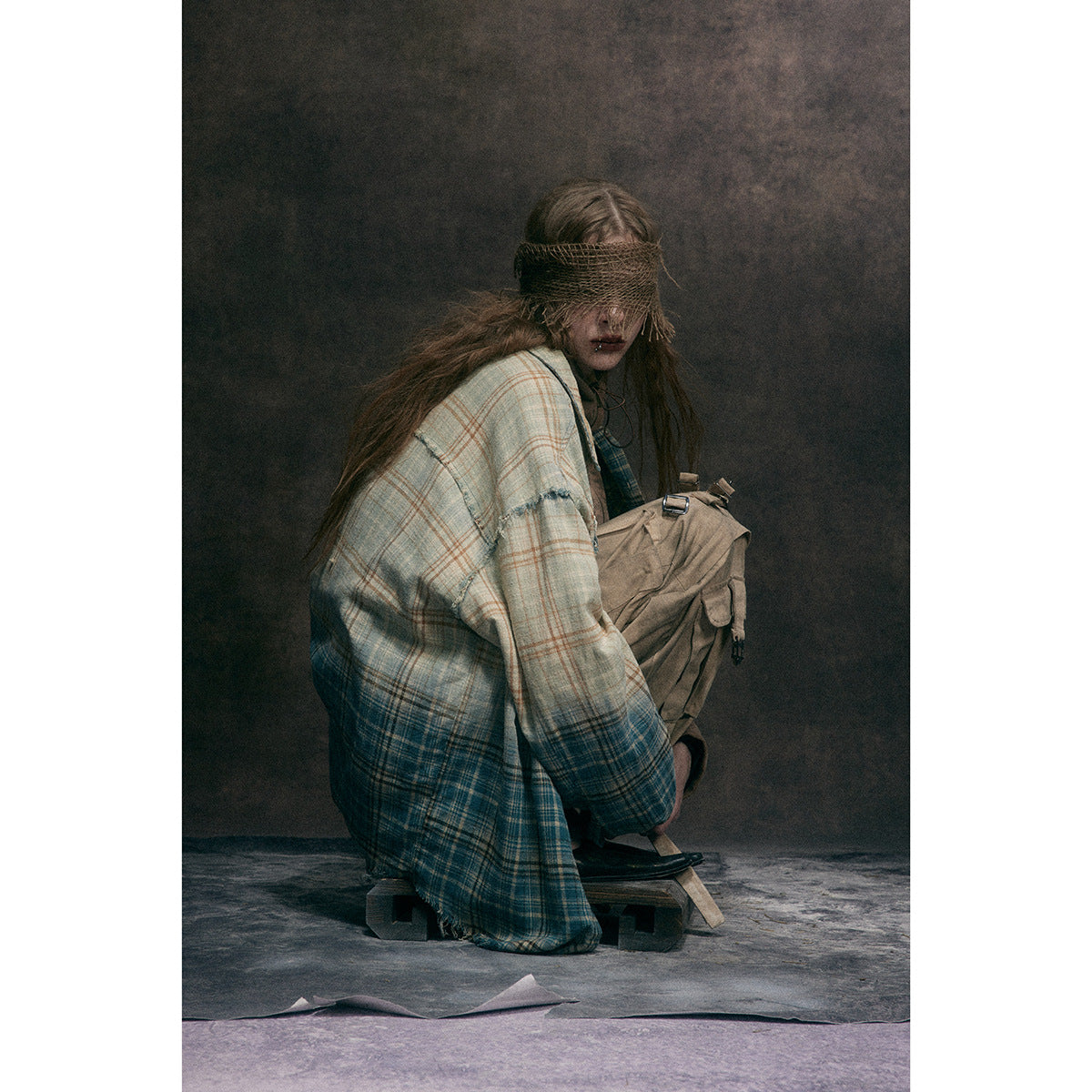 Seated model wearing an oversized cream-and-blue plaid shirt draped over tan cargo trousers.