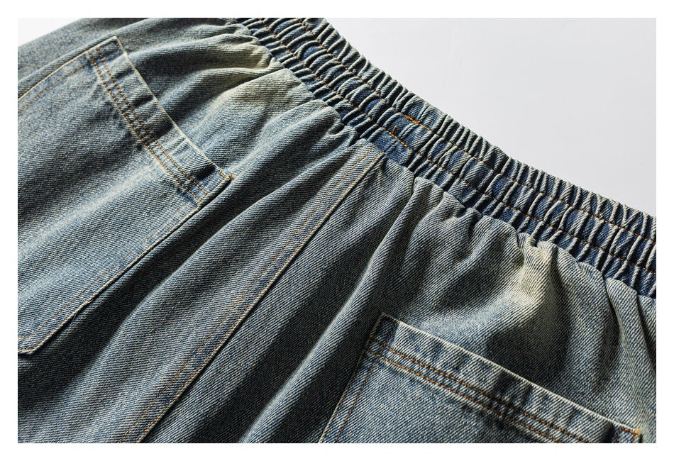 Close-up of washed blue denim pants showing a gathered elastic waistband and two pockets with orange stitching.