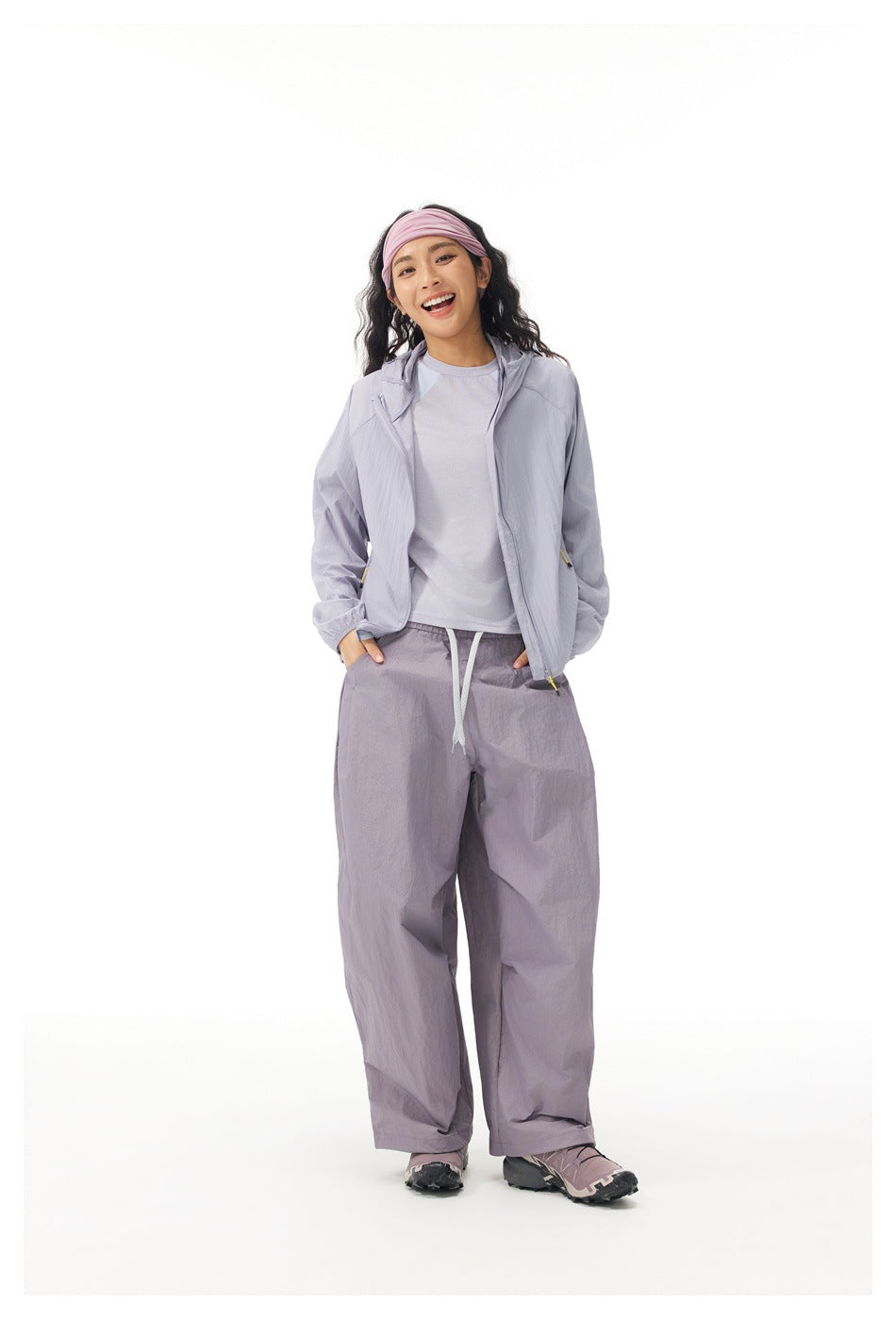 Full-length model wearing an open light gray-lavender hooded jacket over a pale shirt and loose trousers.