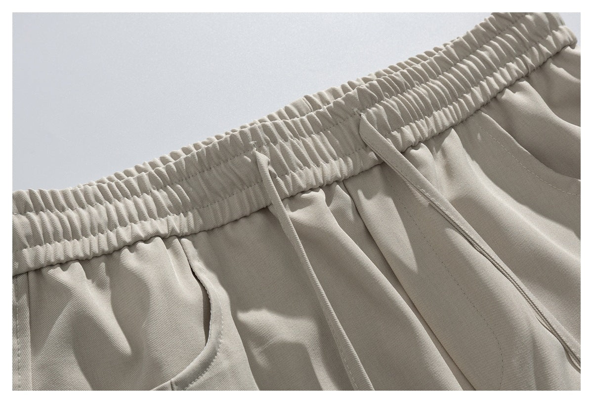 Close-up of beige trousers showing the gathered elastic waistband, drawstrings, and curved pocket opening.