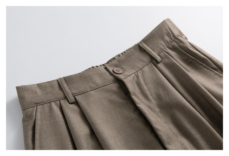 Close-up of taupe shorts showing the waistband, button, belt loops, pleats, and front fly.
