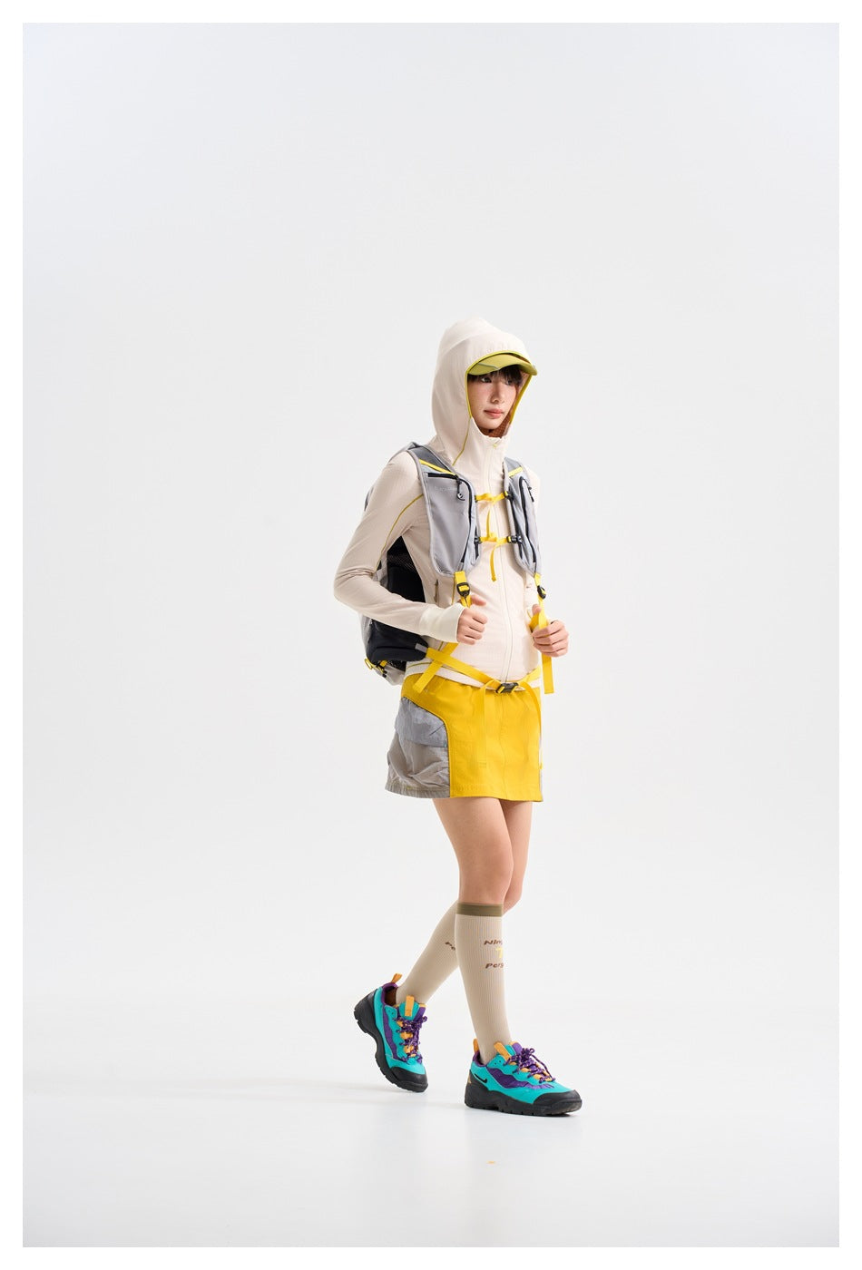 Model wearing a cream hooded jacket with yellow piping, a yellow-and-gray skirt and a gray hiking backpack.