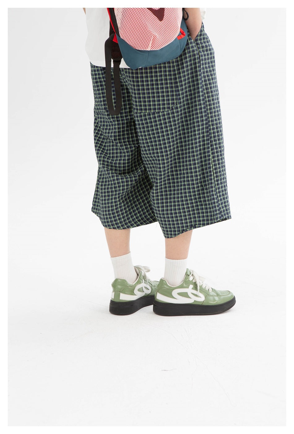 Cropped model wearing loose navy and green checked shorts with a white top, backpack, socks and green sneakers.