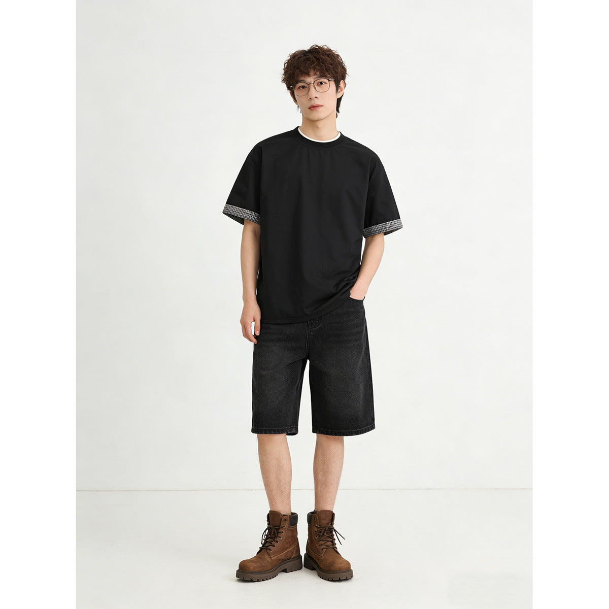 Model wearing a black crew-neck T-shirt with checked sleeve cuffs, black shorts and brown boots.