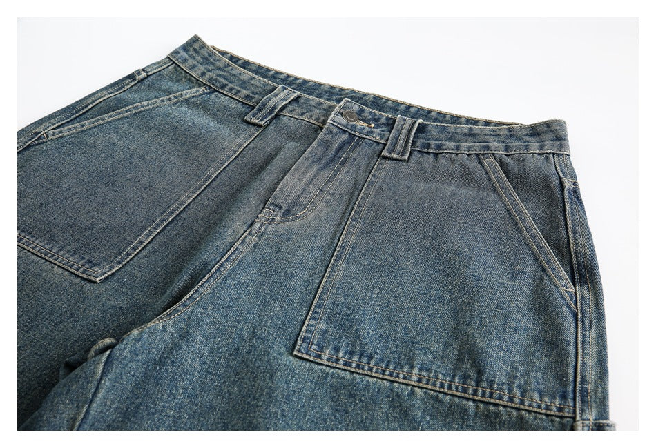 Close view of blue washed denim trousers showing the waistband, belt loops, button, and large stitched pockets.