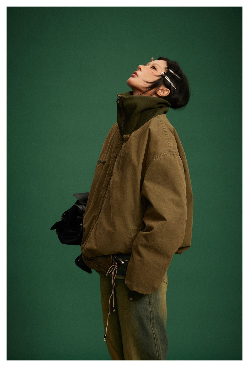 Model wearing an oversized olive-brown jacket with a dark ribbed high collar and front zip, shown in profile.