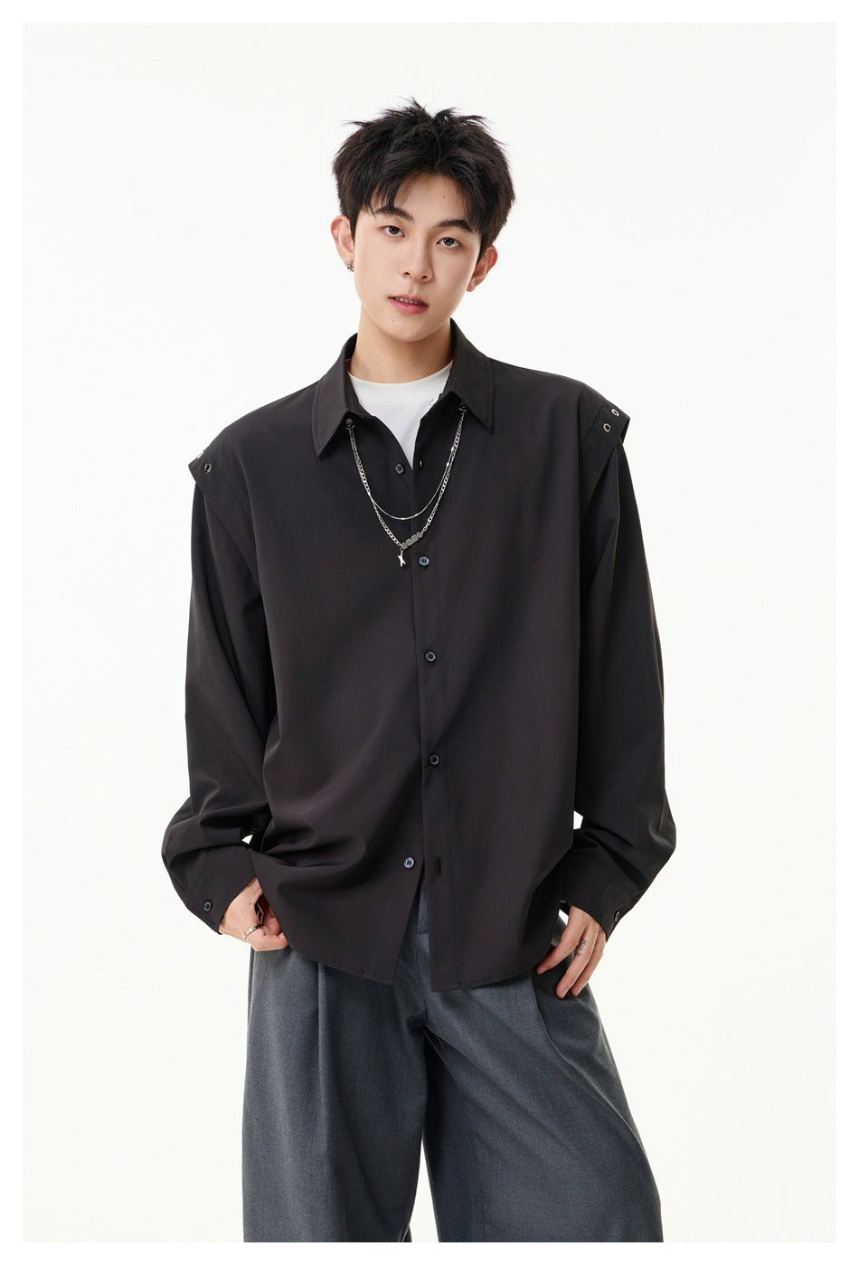 Model wearing an oversized black button-up shirt with shoulder snaps, layered silver chains, and gray wide-leg trousers.