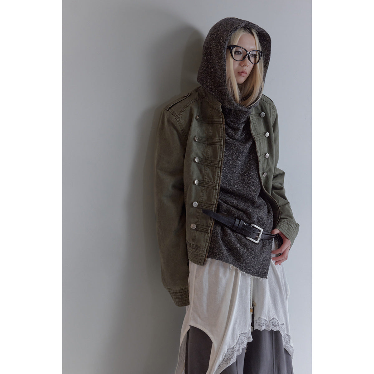 Model wearing an olive military-style buttoned jacket over a charcoal hooded top and layered skirts.