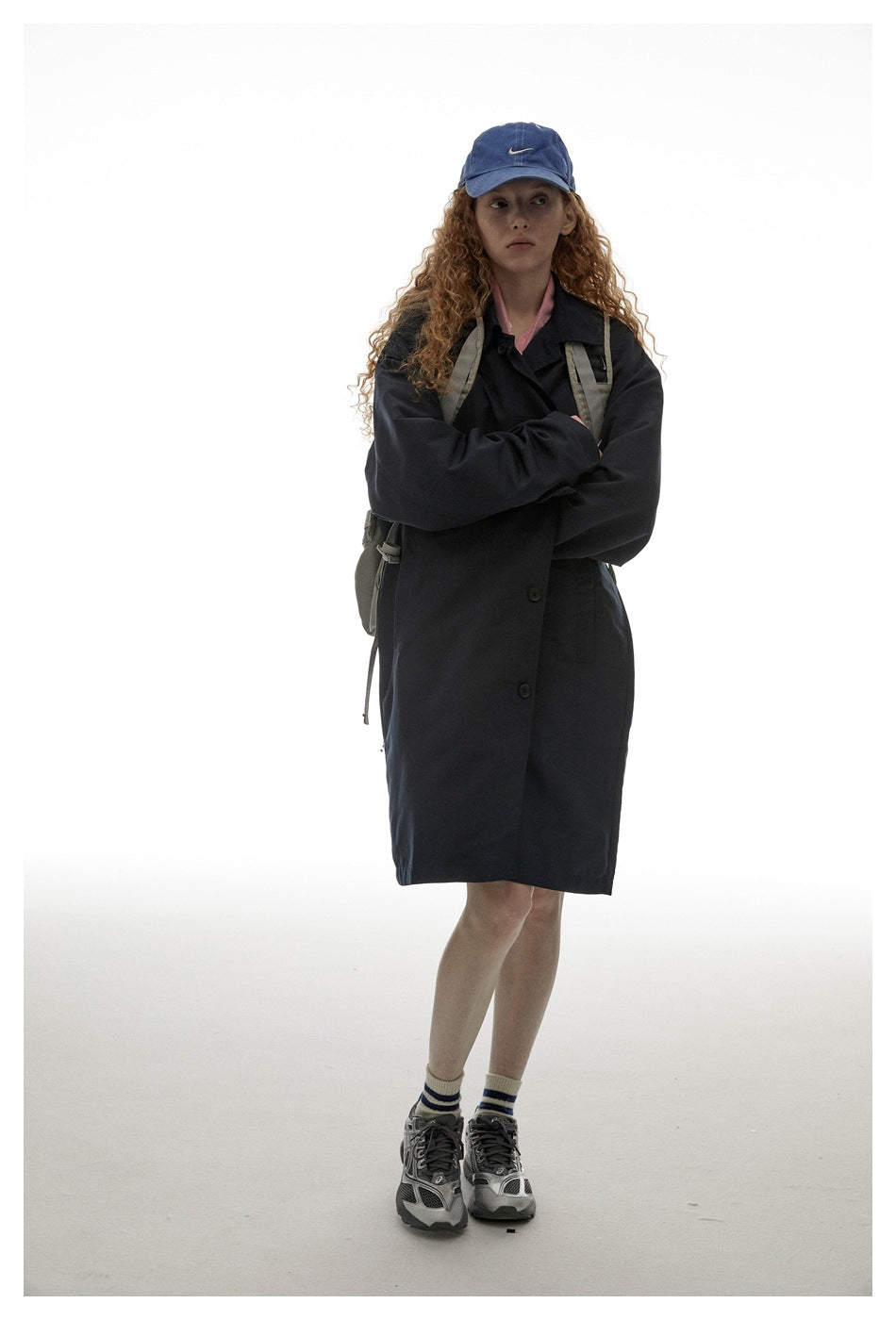 Full-length model wearing a dark navy button-front coat, blue cap, backpack, striped socks and sneakers.