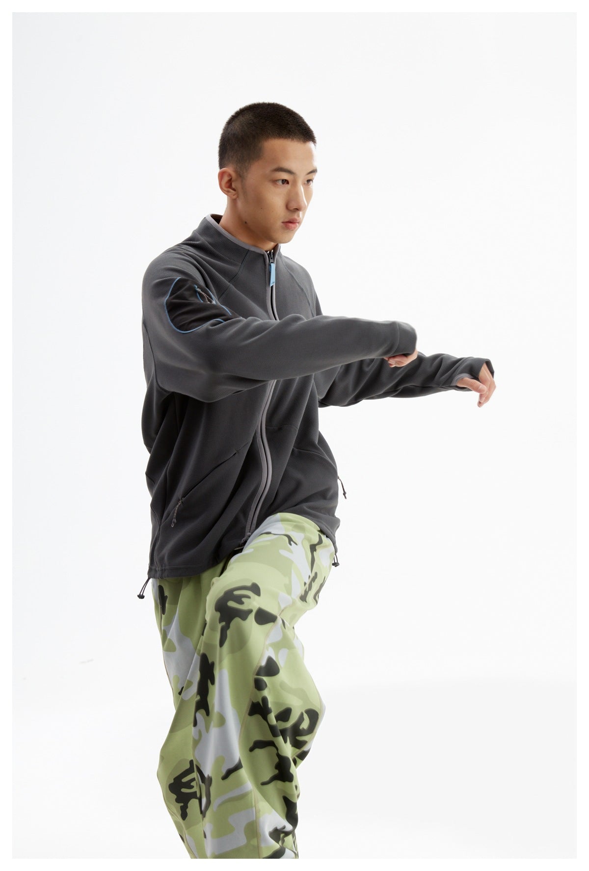 Man modeling a charcoal zip jacket and green camouflage pants while stretching his arms.