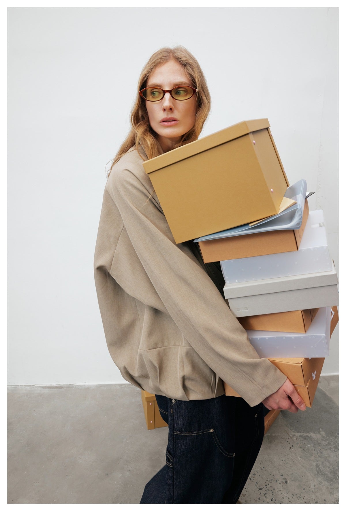 Model in a loose taupe jacket and dark jeans, carrying a stack of boxes that obscures much of the jacket.