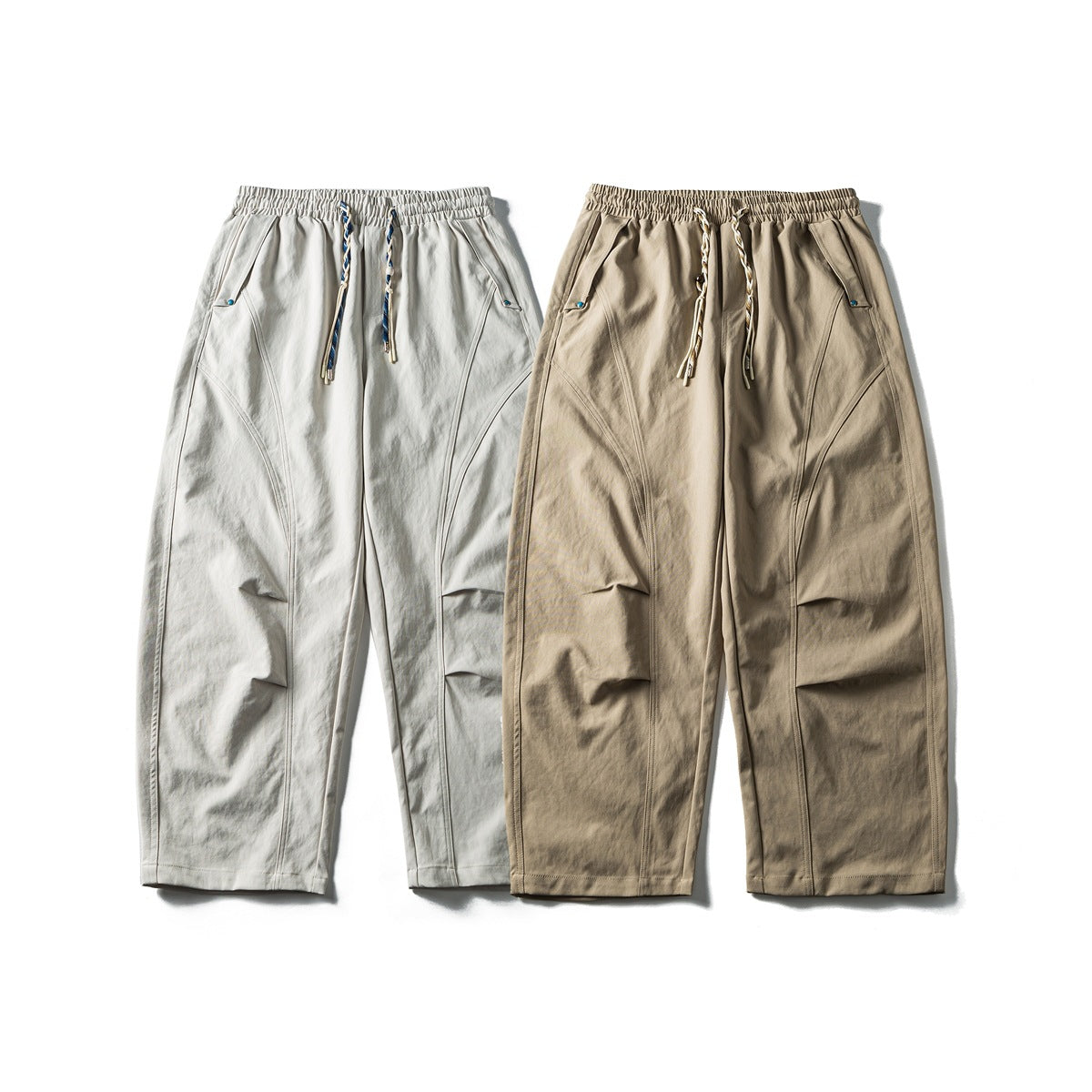 Front view of loose trousers in light gray and taupe, with elastic waists, braided drawstrings, and curved seam panels.