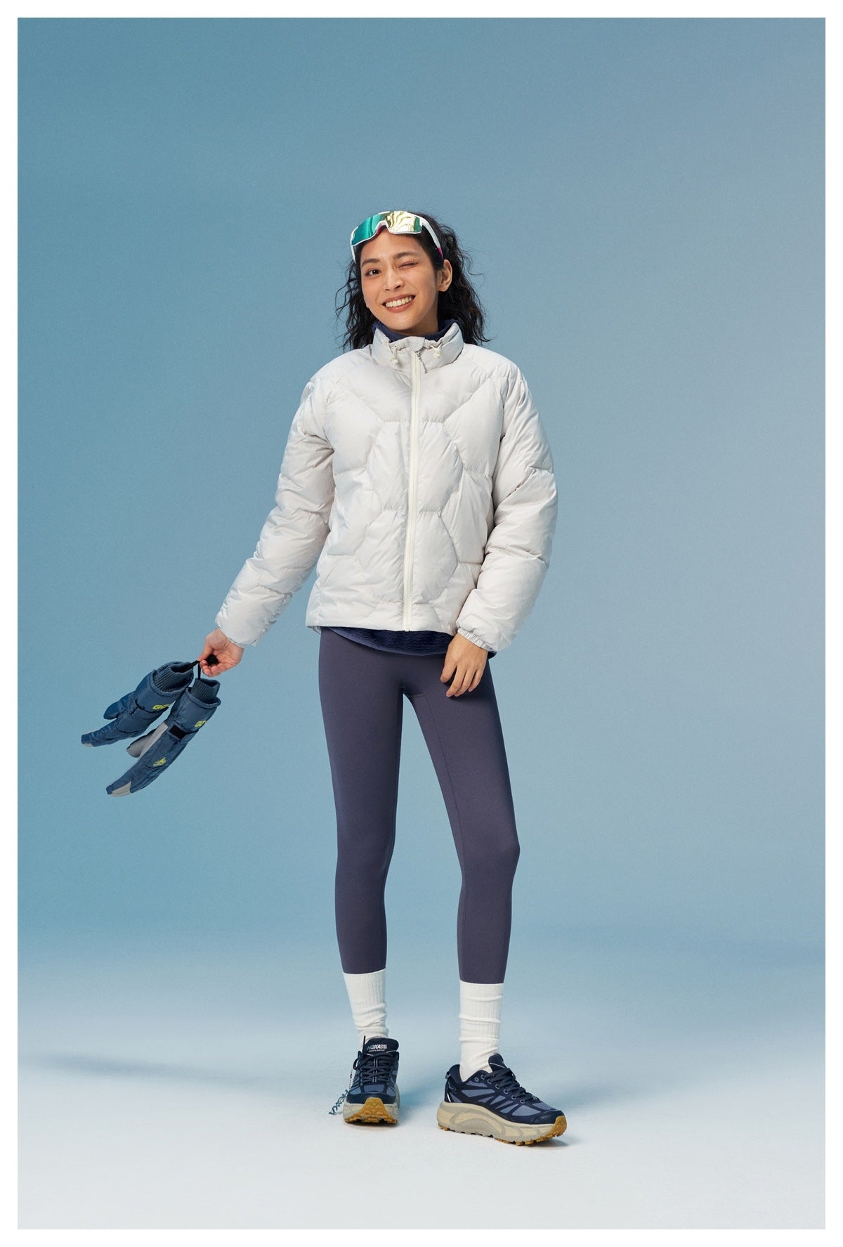 Woman wearing a white quilted puffer jacket and dark leggings, holding a pair of blue shoes.