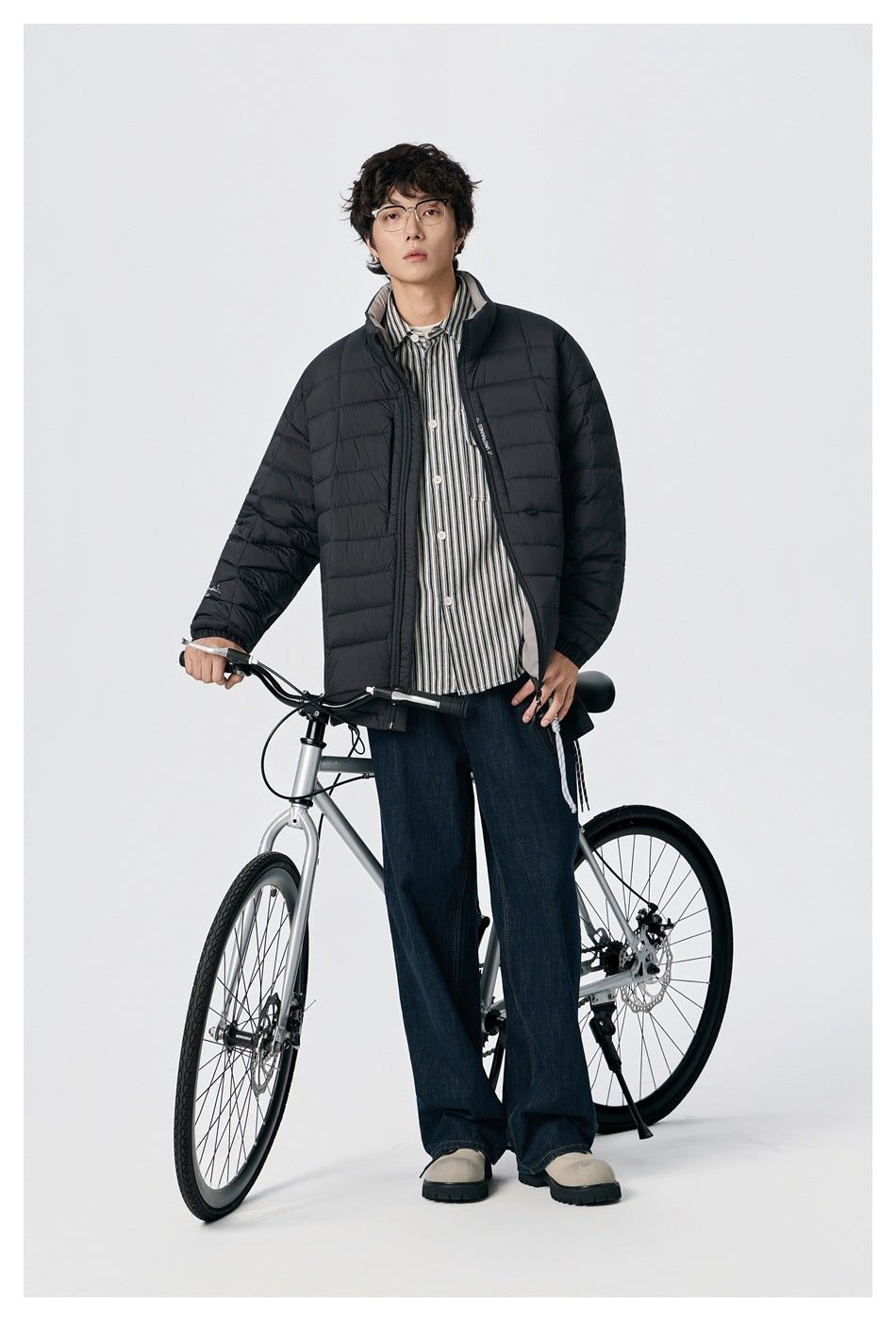 Model wearing an open black quilted jacket beside a bicycle, shown nearly full length.