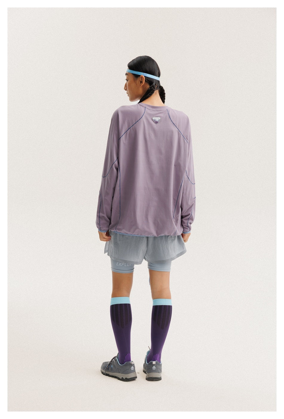 Back view of a woman wearing a lavender long-sleeve top with blue seam piping, gray running shorts, and purple knee socks.