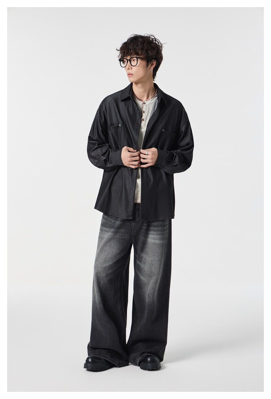Model in a dark charcoal two-pocket button-up shirt and faded wide-leg jeans, standing with hands at the shirt front.