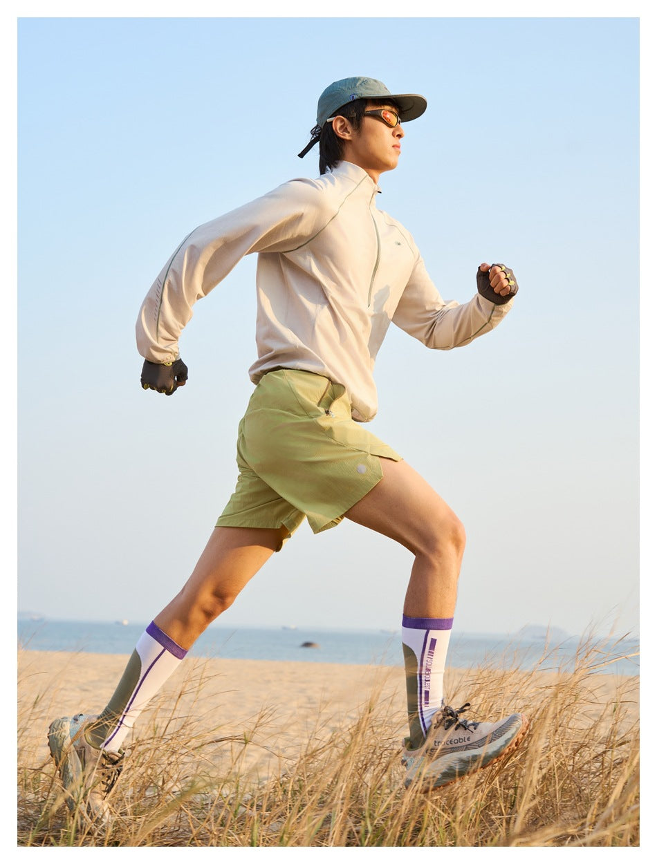 Man running on a beach in a pale beige quarter-zip top, olive shorts, cap, gloves and tall socks.