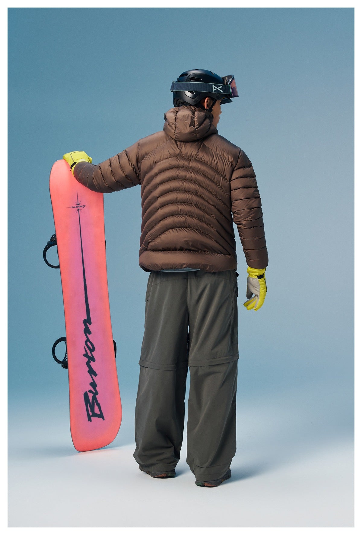 Rear view of a model wearing a dark brown quilted hooded jacket beside a pink snowboard.
