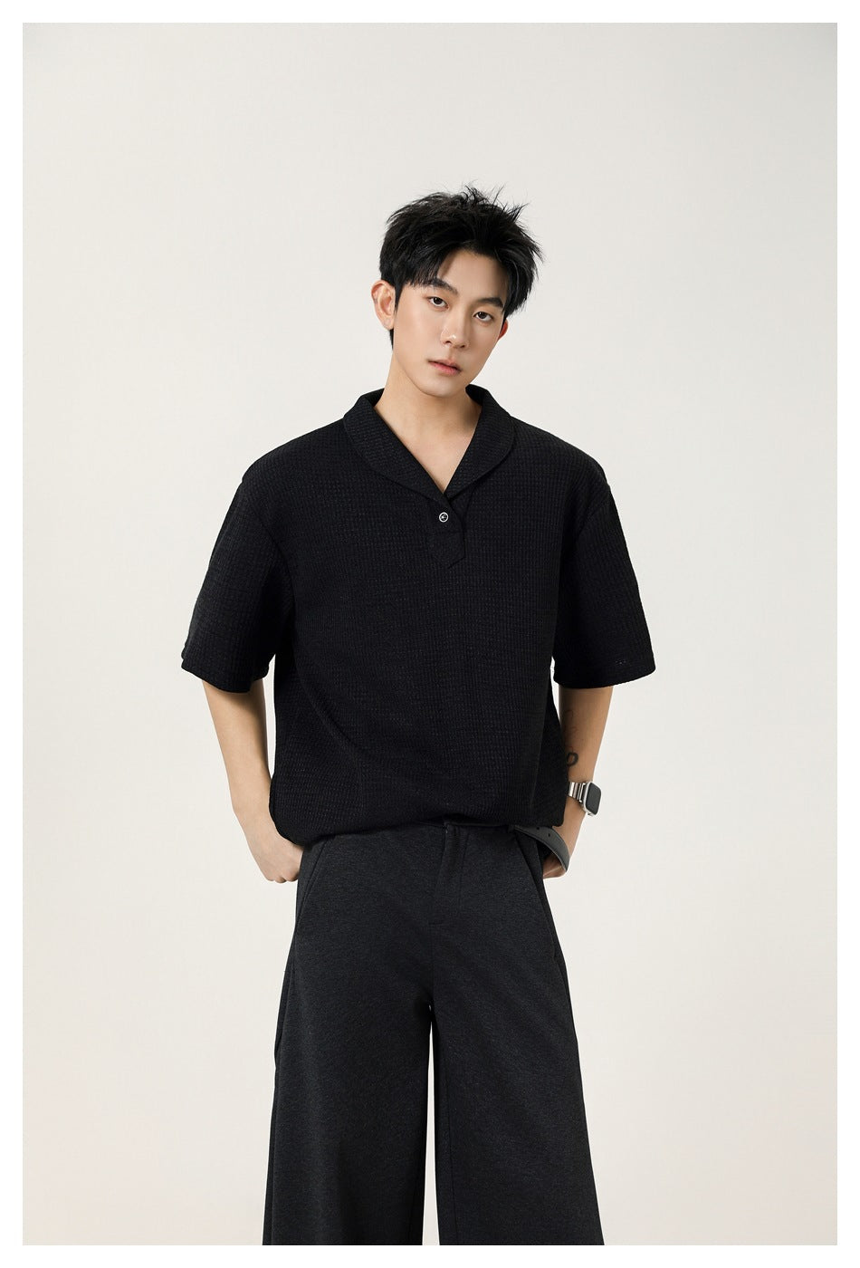 Model wearing a black short-sleeve textured polo with a notched shawl collar and dark wide-leg trousers.