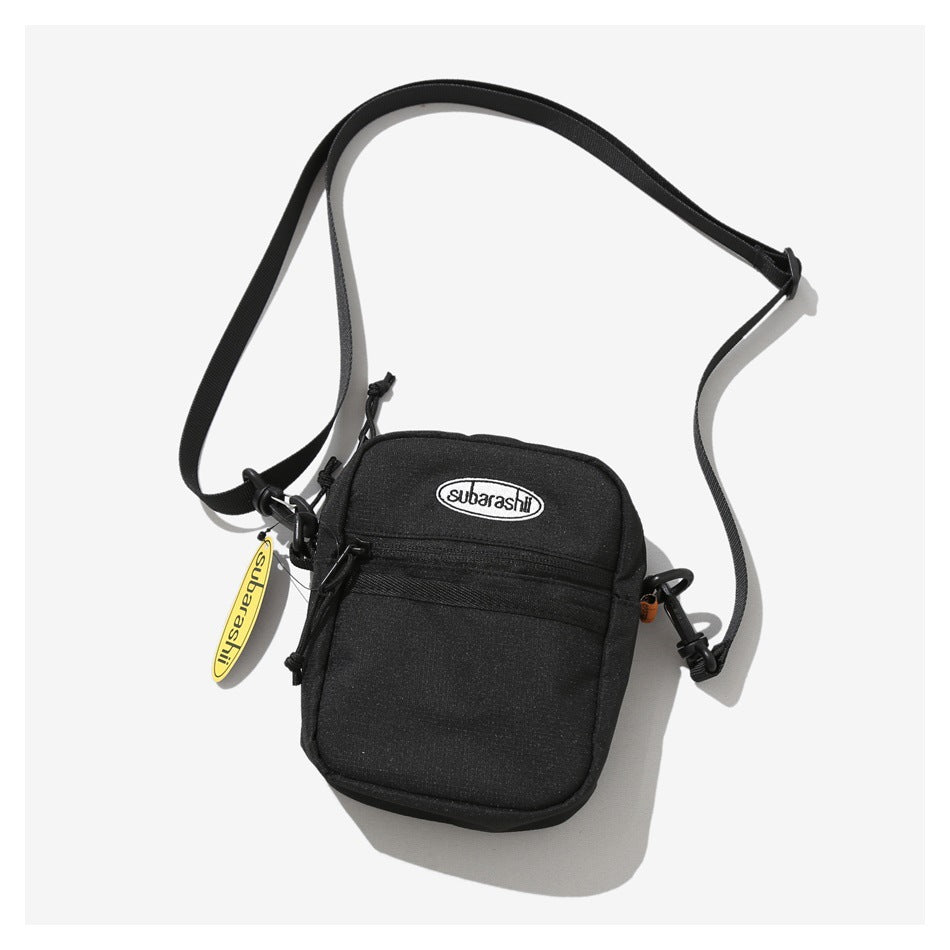 Black crossbody bag with front zip pocket, oval logo patch, black strap and yellow tag on a light background