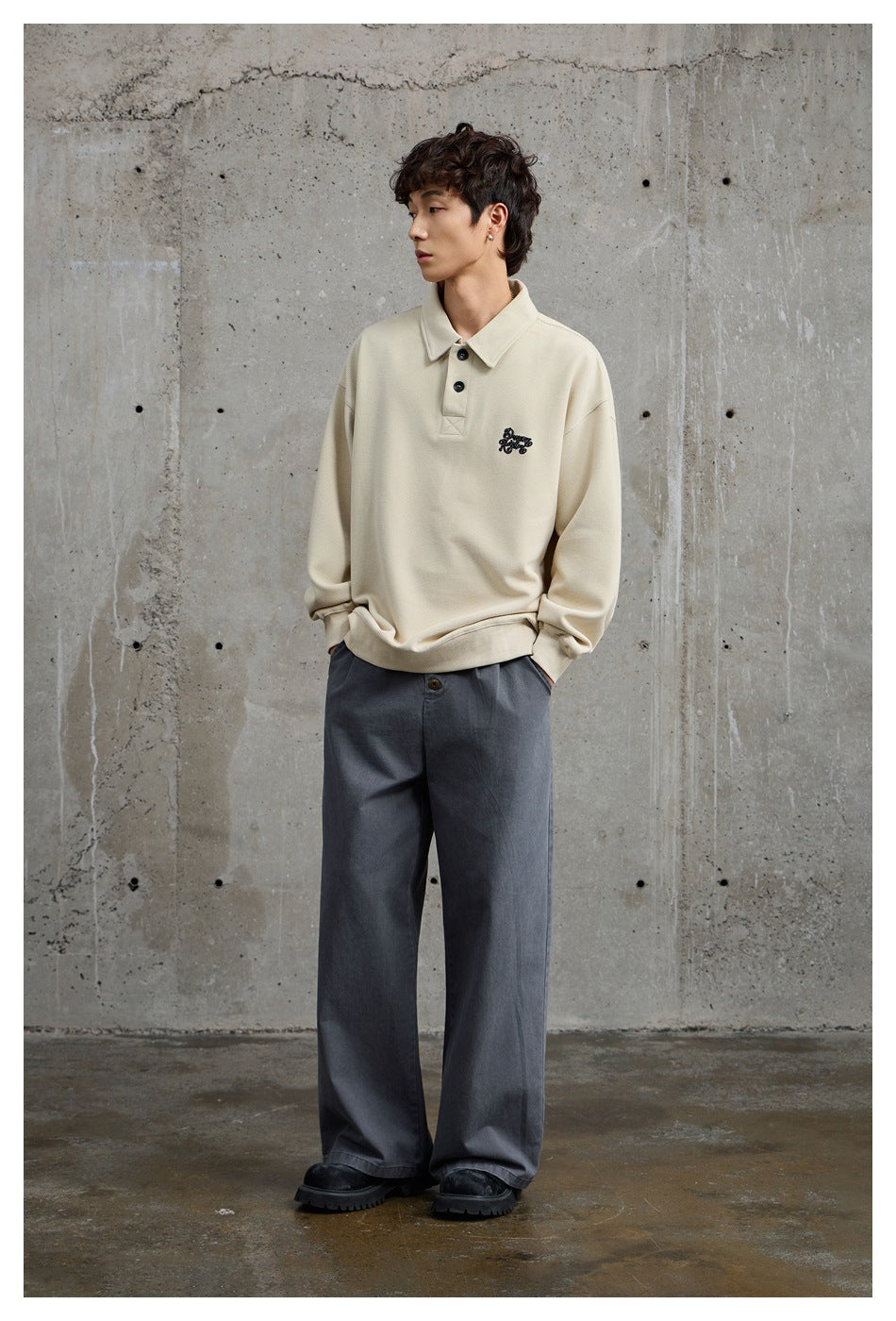 Full-length view of a model wearing a light beige collared pullover with chest embroidery and wide gray trousers.