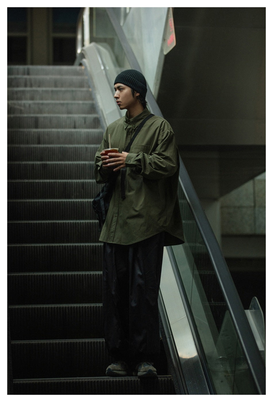 Model wearing an oversized olive-green button-up shirt with a chest pocket and black trousers.