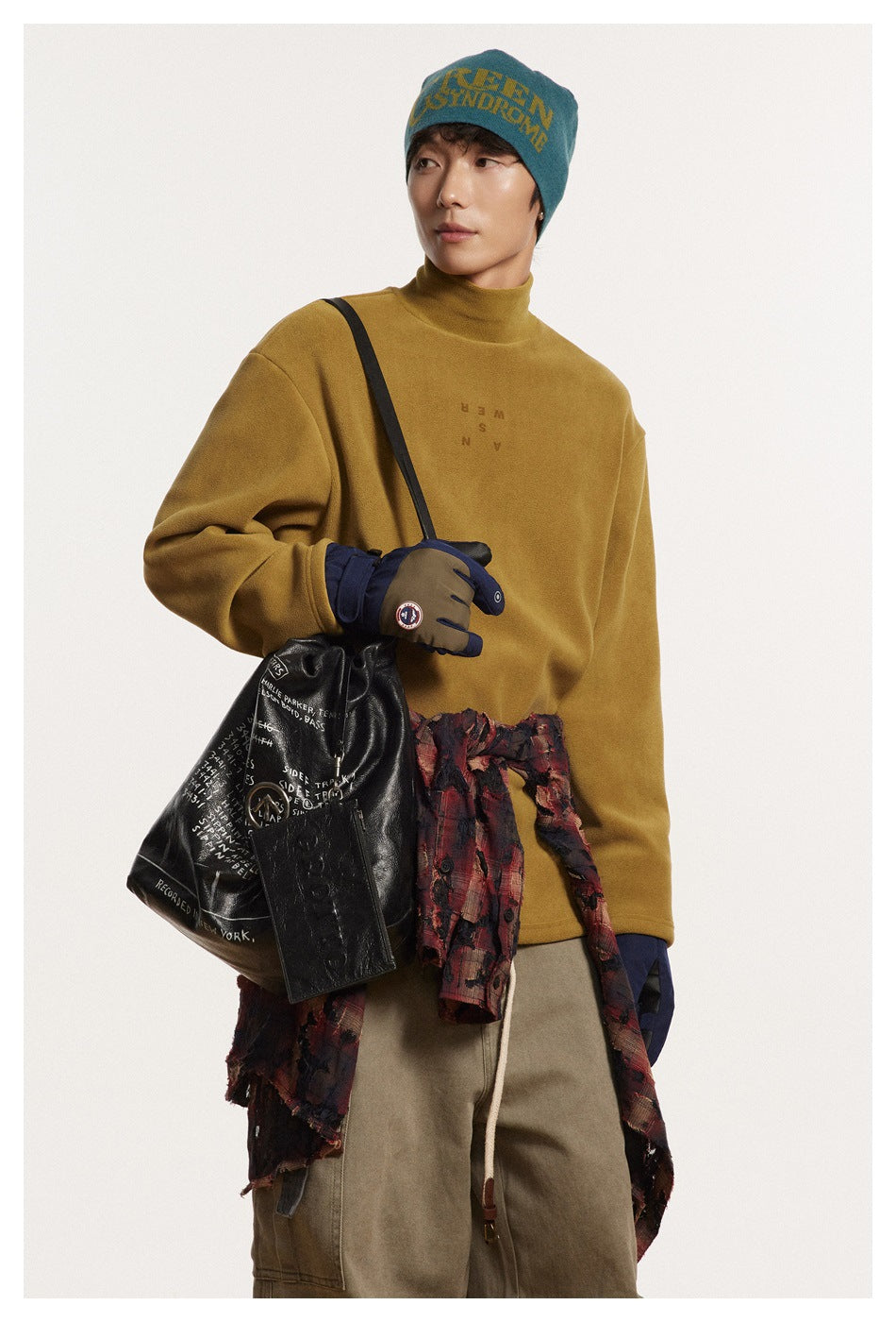 Model in a mustard high-neck pullover and teal beanie, carrying a black bag with a plaid shirt tied at the waist.