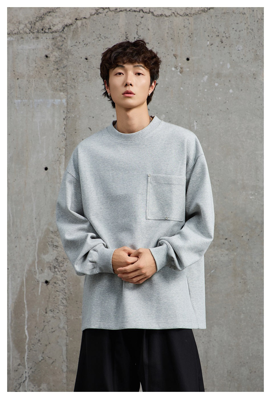 Model wearing a light gray oversized crewneck sweatshirt with a snap-detail chest pocket and black trousers.