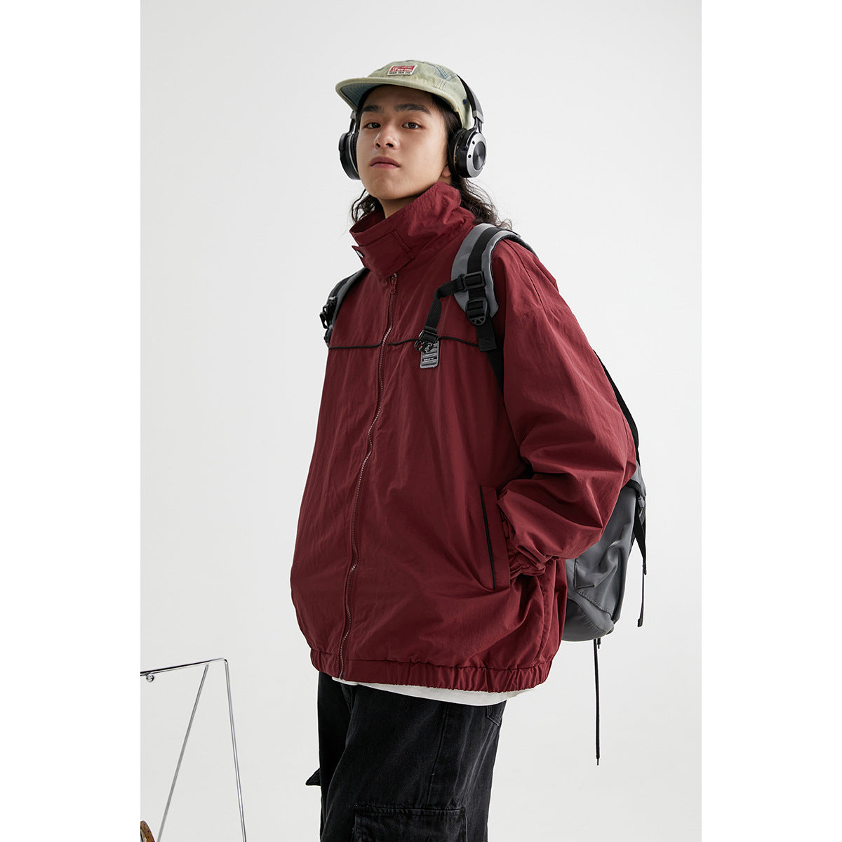 Model wearing a burgundy zip jacket with a high collar, black piping and a chest patch; backpack straps are visible.