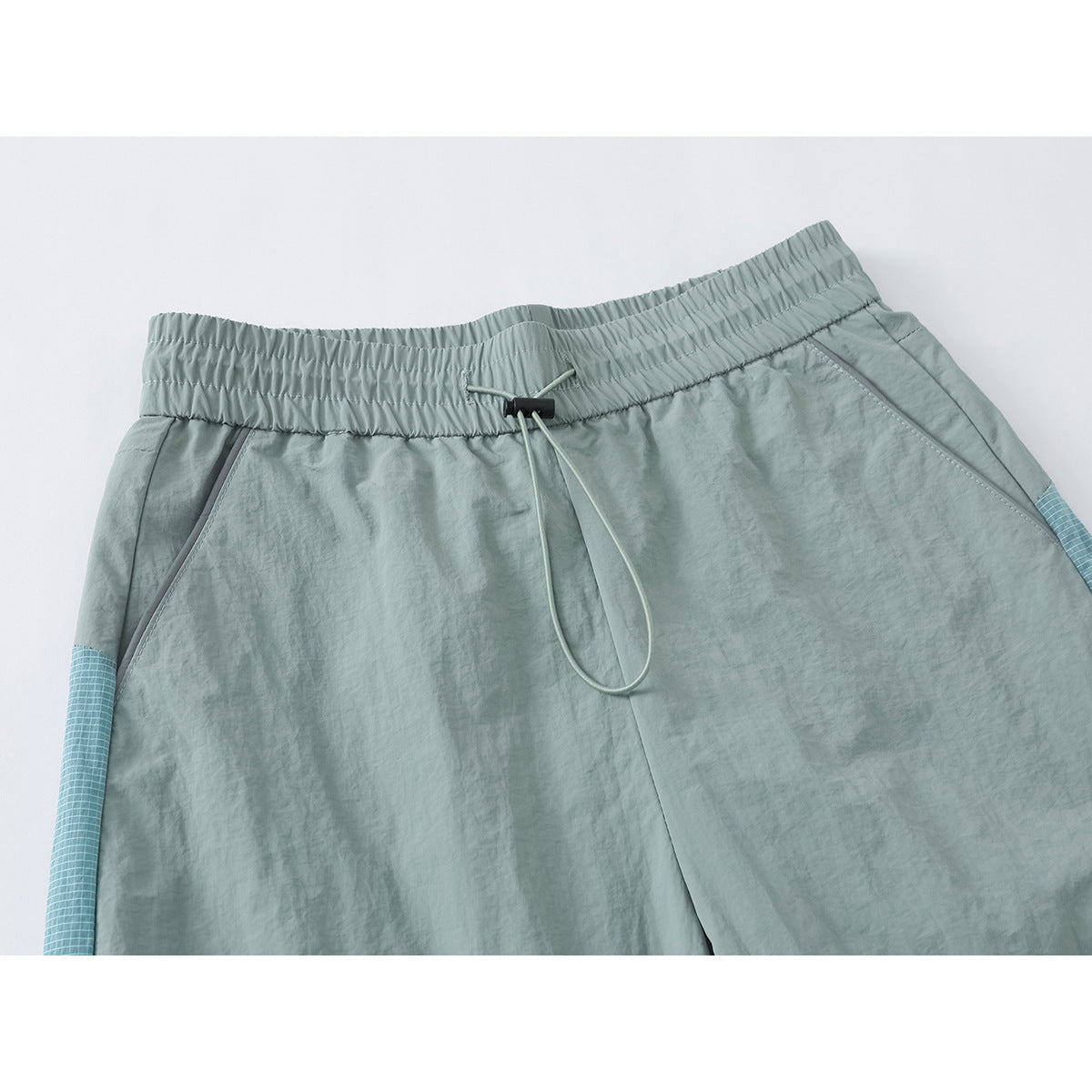 Close-up of pale sage wide-leg pants showing the gathered elastic waist, drawcord, slanted pockets and blue grid side panels.