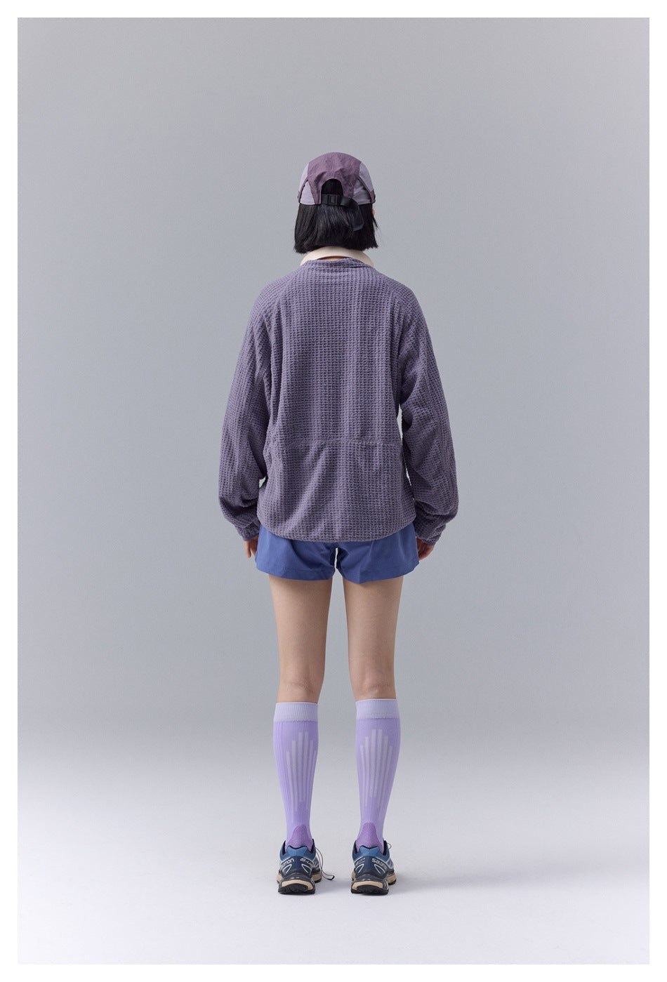 Back view of a model wearing a textured lavender jacket, blue shorts, and a lavender cap.