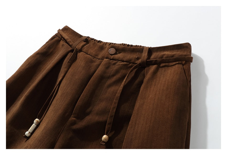 Cropped front view of brown trousers showing the buttoned gathered waist, pleats and beaded drawcords.