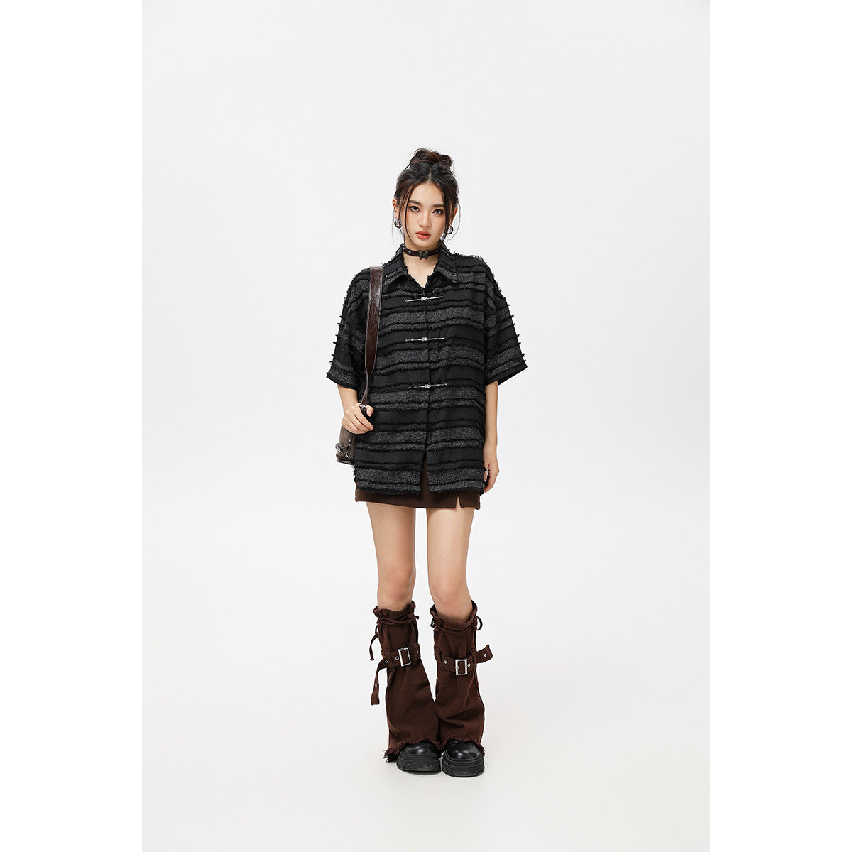Model wearing an oversized black striped short-sleeve shirt with metal clasps, a brown mini skirt, and tall brown boots.