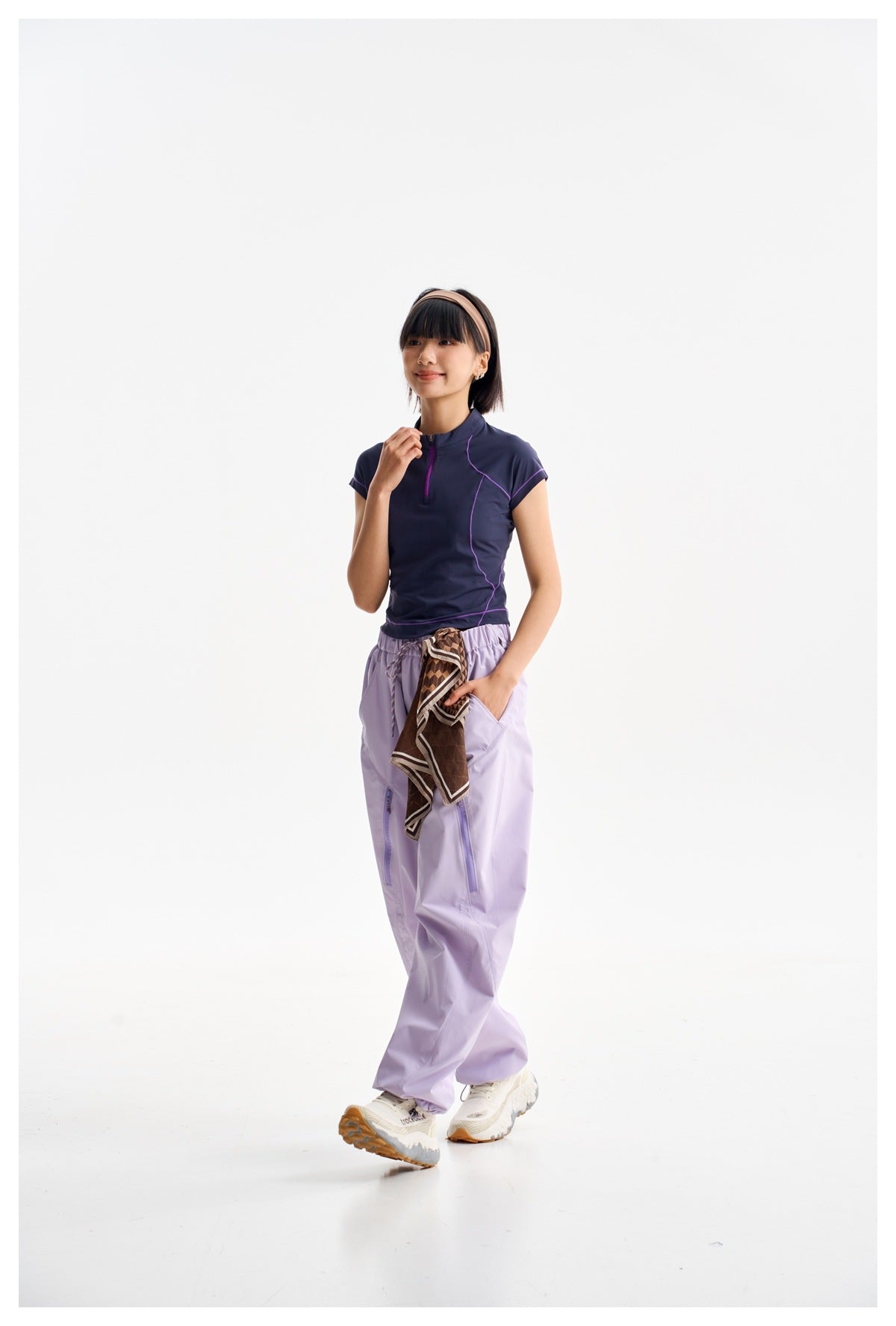Model wearing a navy short-sleeve zip-neck top with purple piping and loose lavender pants, walking in white sneakers.