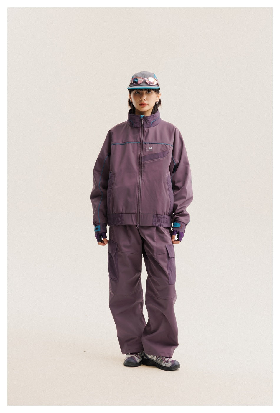 Full-length front view of a model wearing a plum zip jacket with turquoise piping and matching cargo pants.