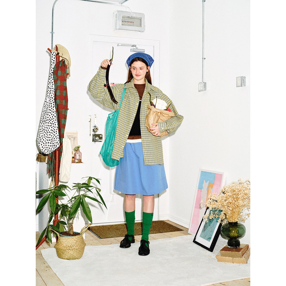 Model in a yellow-and-blue plaid shirt and blue skirt, holding a paper bag and raising a long dark vegetable.