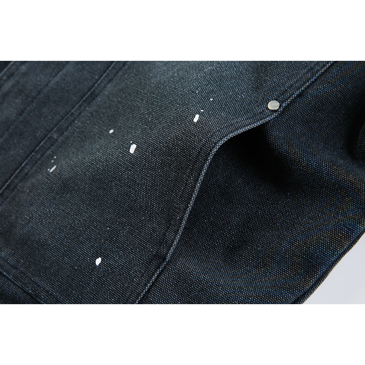 Close-up of dark textured jacket fabric with scattered white paint-like marks and a silver snap.