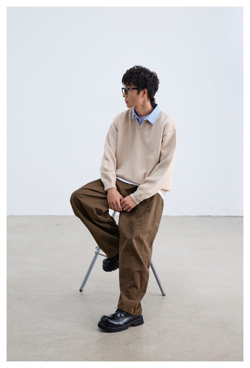 Seated model wearing a beige V-notch sweater over a blue striped shirt with loose brown trousers.