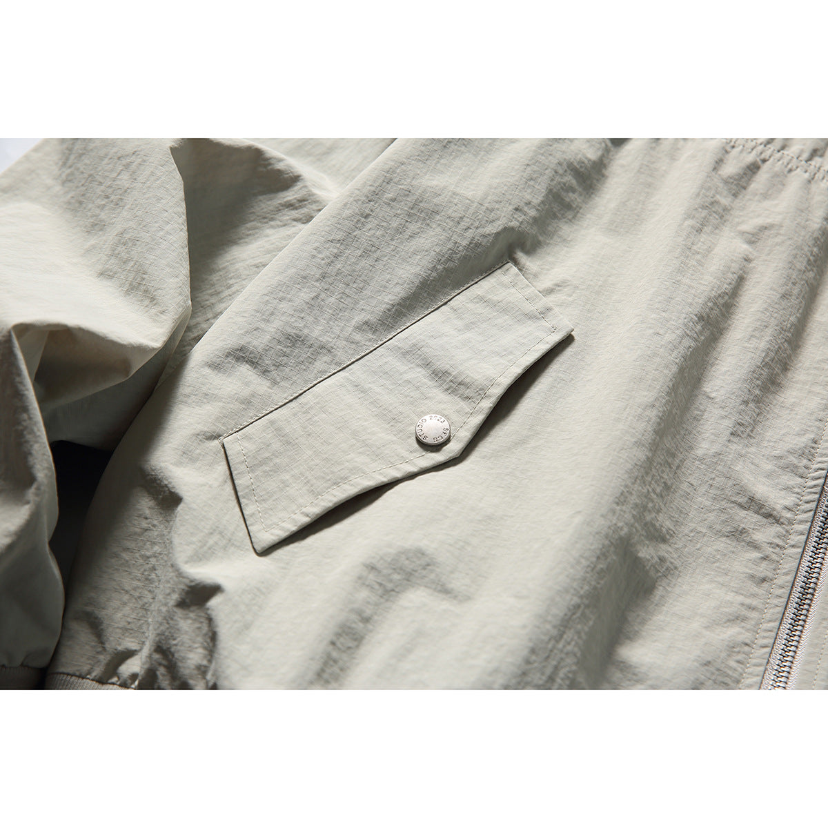 Close-up of a light gray-beige jacket's angled flap pocket with a snap button and front zipper edge.