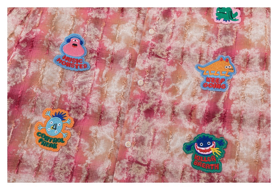 Close-up of the shirt front with colorful embroidered monster patches and buttons on distressed plaid fabric.