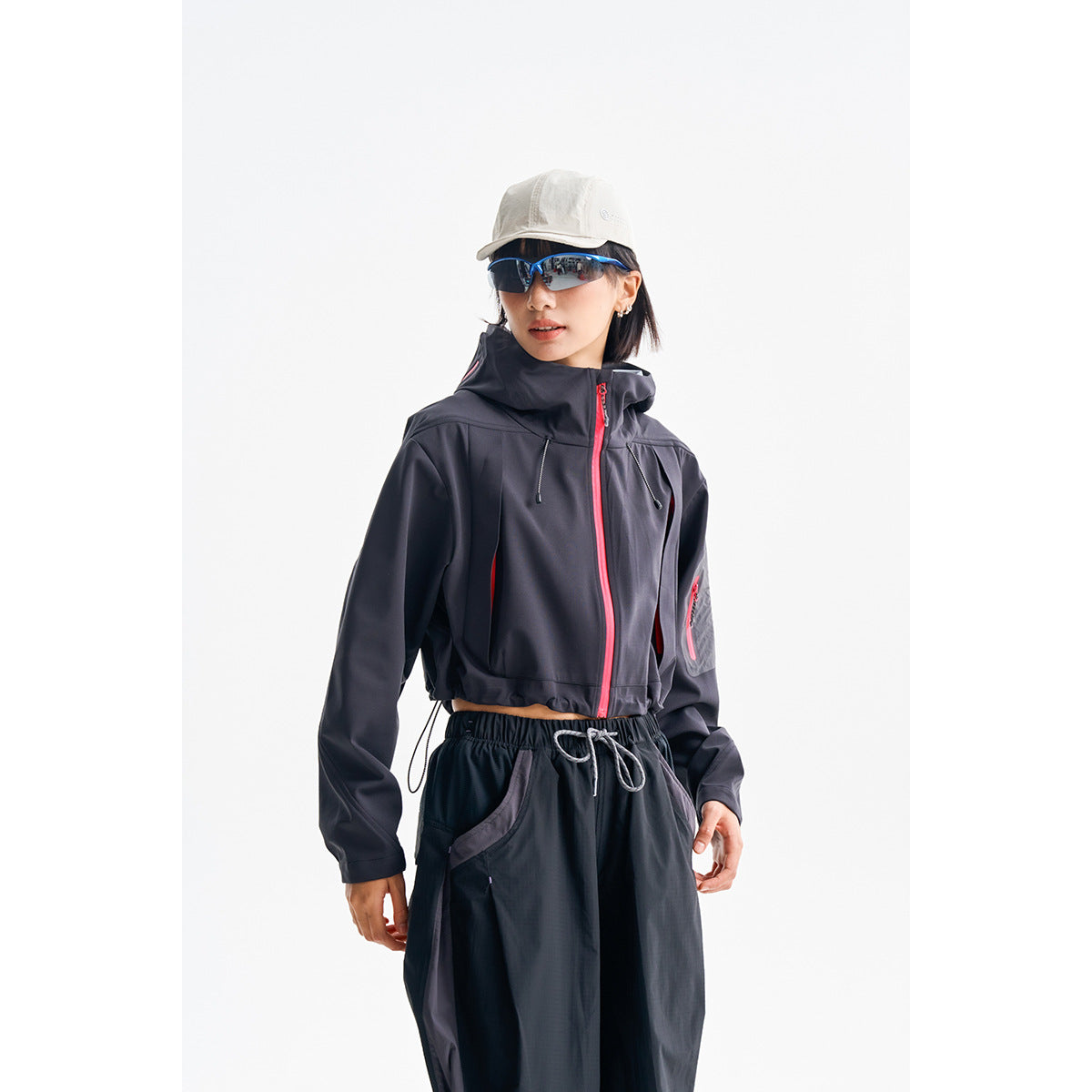 Model wearing a dark cropped hooded jacket with a bright pink front zipper and matching sleeve pocket zipper.