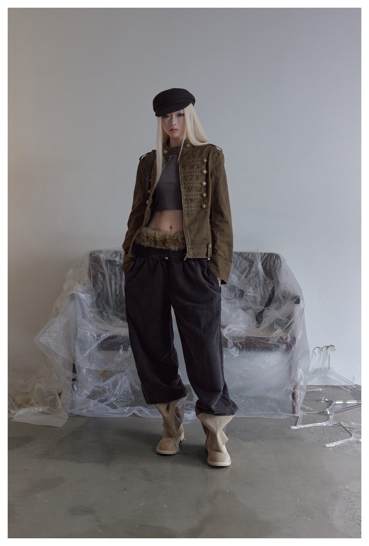 Model wearing the brown button-trim short jacket open over a cropped top, dark pants and a black cap