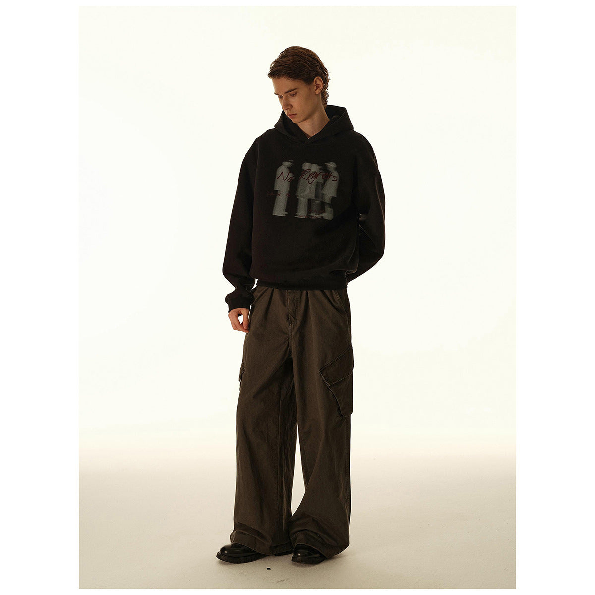 Full-length view of a model in a black “No Regrets” graphic hoodie and wide dark cargo trousers.