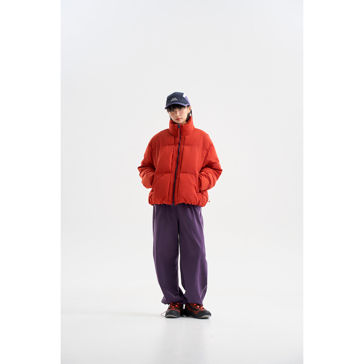 Model wearing a bright red high-collar puffer jacket with dark purple trousers and a navy cap.